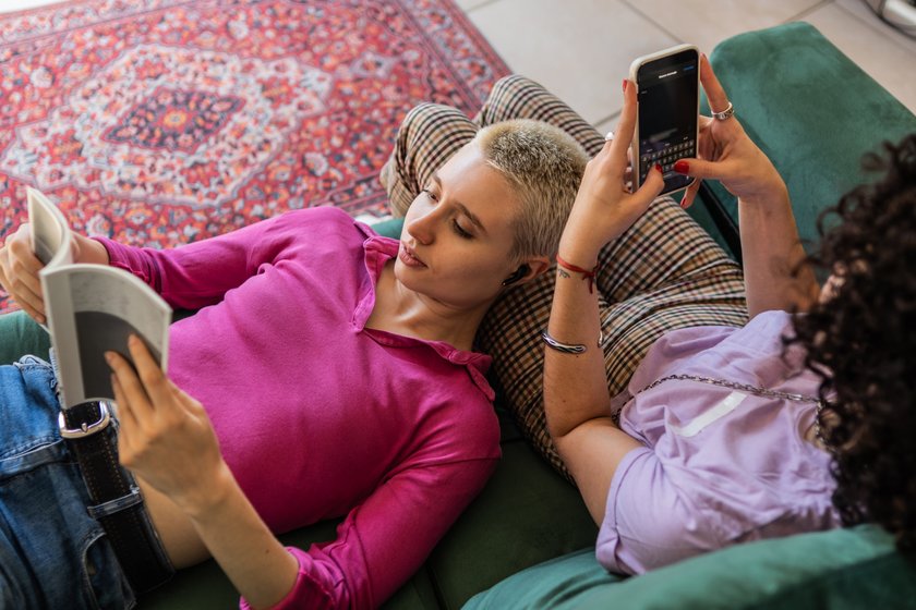 Young woman reading a book lying on her friend's lap on the sofa at home Young woman reading a book lying on her friend's lap on the sofa at home