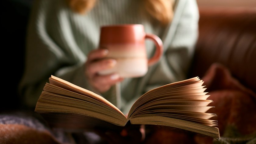 Eine Frau hält in der einen Hand ein aufgeschlagenes Buch und in der anderen eine Tasse Eine Frau hält in der einen Hand ein aufgeschlagenes Buch und in der anderen eine Tasse