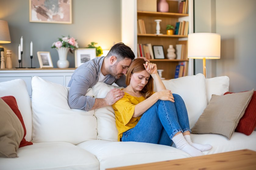 Er sagt nicht "Ich liebe dich": Frau sitzt traurig auf der Couch, während ihr Partner sie tröstet Er sagt nicht "Ich liebe dich": Frau sitzt traurig auf der Couch, während ihr Partner sie tröstet