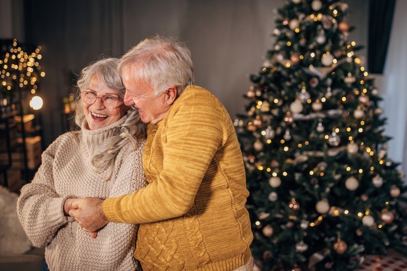 Boomer-Adventskalender: Älteres Paar liegt sich vor einem Weihnachtsbaum in den Armen