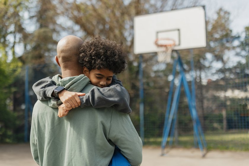 Sätze, die dein Kind zum Reden bringen: Papa und Sohn umarmen sich auf Sportplatz Sätze, die dein Kind zum Reden bringen: Papa und Sohn umarmen sich auf Sportplatz