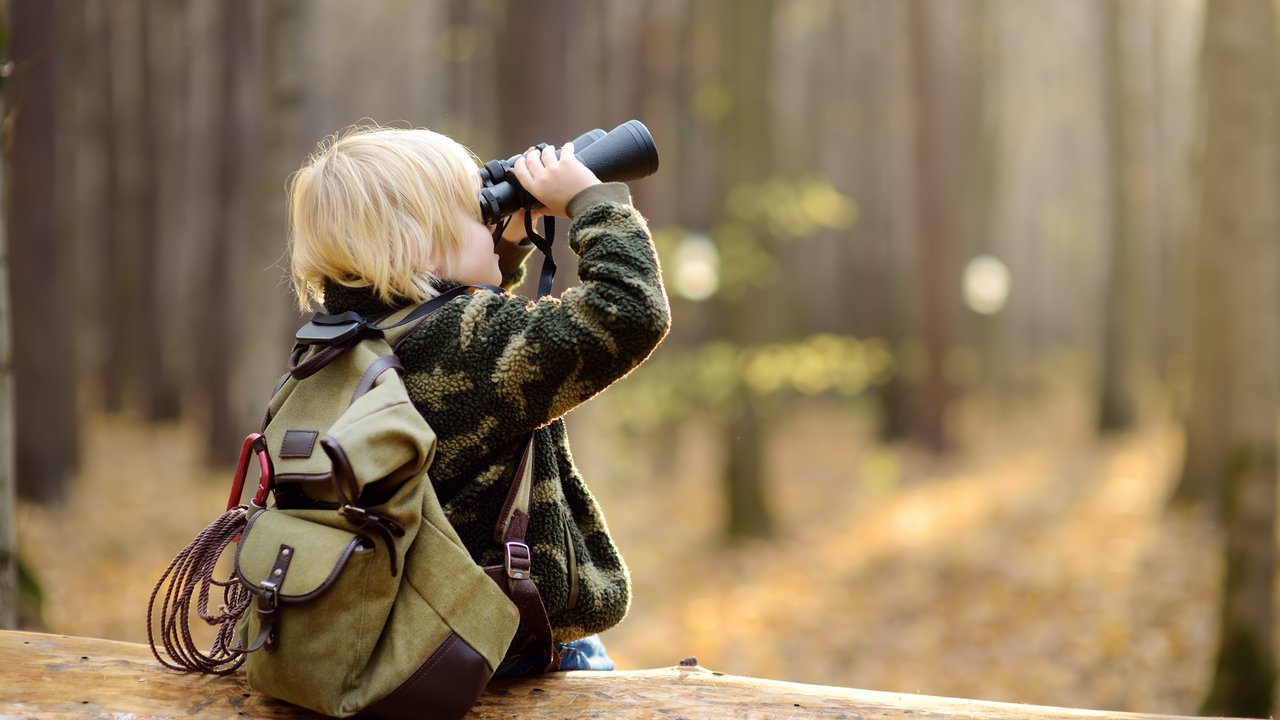 Little boy scout with binoculars during hiking in autumn forest. Child is sitting on large fallen tree and looking through a binoculars. Concepts of adventure, scouting and hiking tourism for kids.