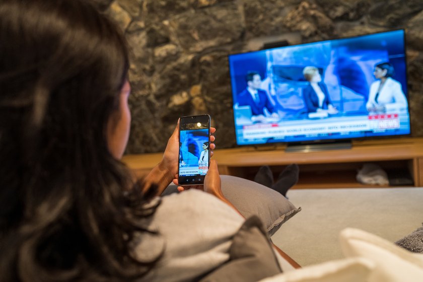 Junge Frau schaut gleichzeitig Nachrichten im Fernsehen und auf dem Smartphone. Junge Frau schaut gleichzeitig Nachrichten im Fernsehen und auf dem Smartphone.