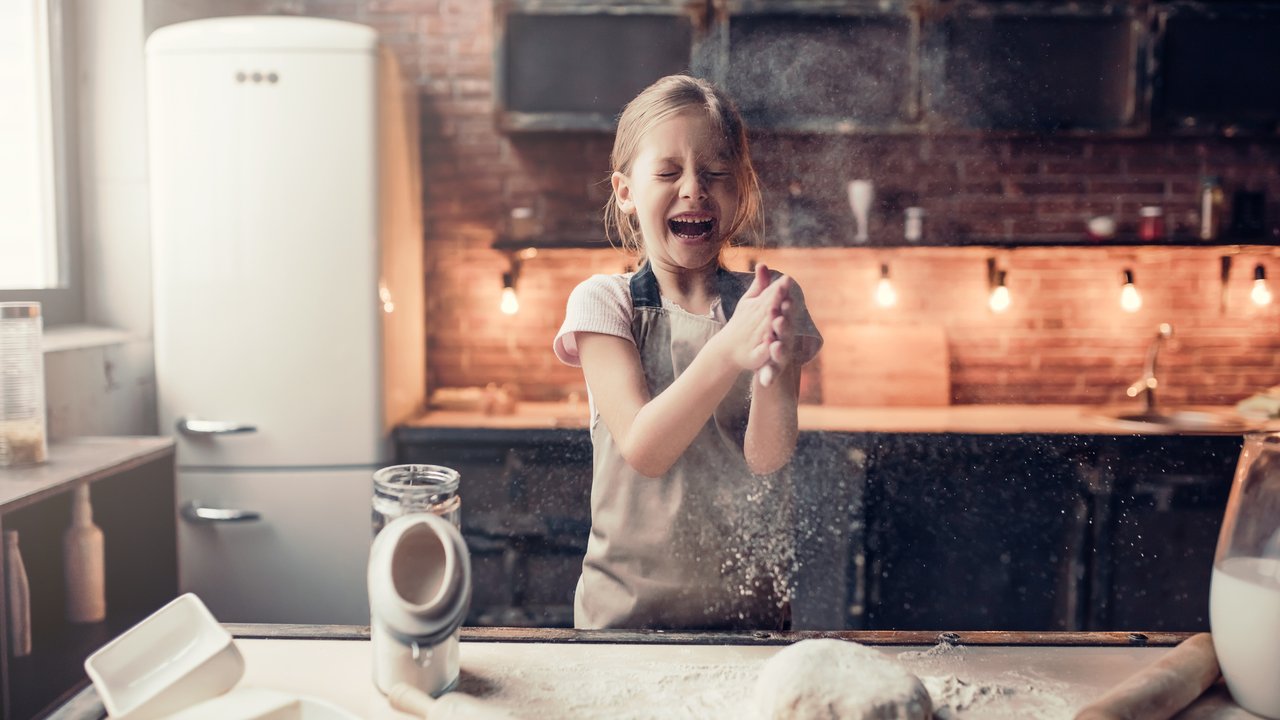 Little cute girl on kitchen is ready for cooking. Laughing while clapping hands with flour.