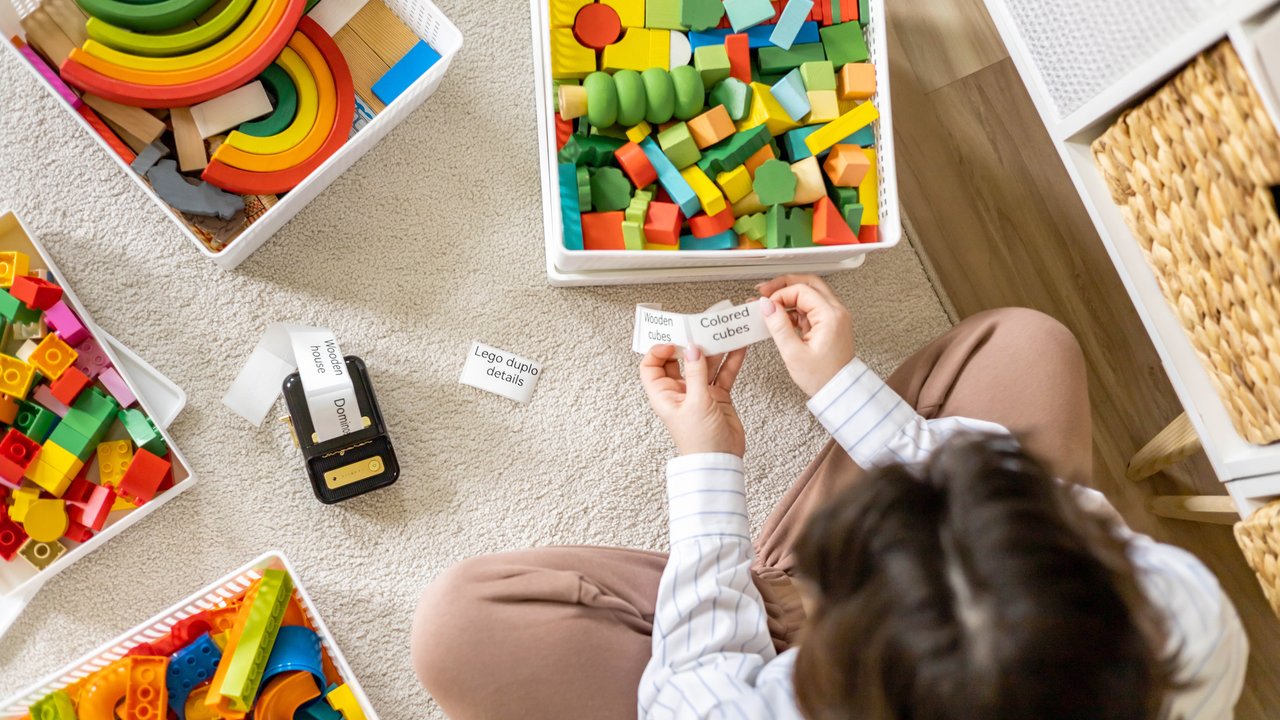 Woman clean out childish room sorting wooden and plastic toys use mobile label printer for sticker with title. Female housewife organizing comfortable storage for plaything with sticky text technology Woman clean out childish room sorting wooden and plastic toys use mobile label printer for sticker with title. Female housewife organizing comfortable storage for plaything with sticky text technology