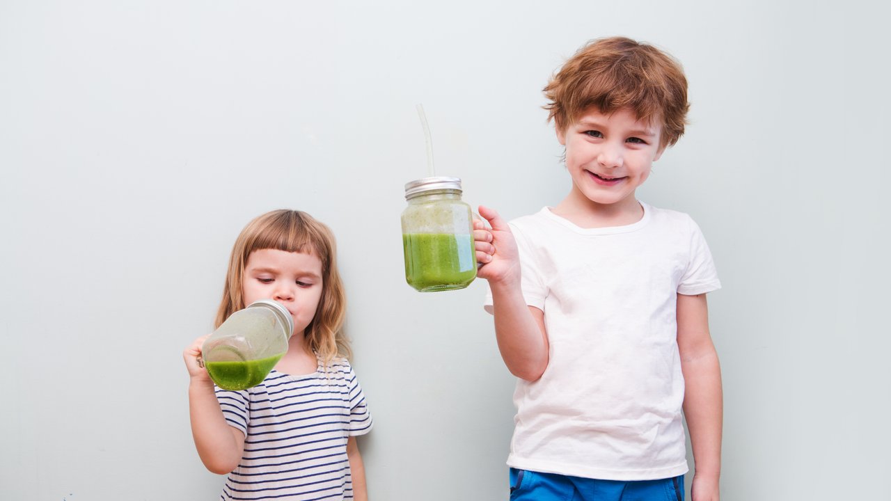 Two Cute smiling kids drinks healthy green smoothie with straw in a jar mug on a gray background