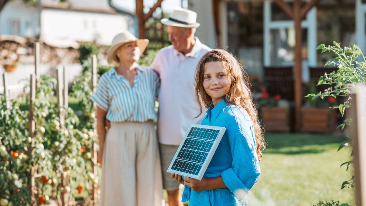 Eine Person in einem blauen Hemd hält ein kleines Solarpanel in einem Garten. Im Hintergrund sind zwei weitere Personen zu sehen, die vor einer Terrasse mit Holzkonstruktion stehen.