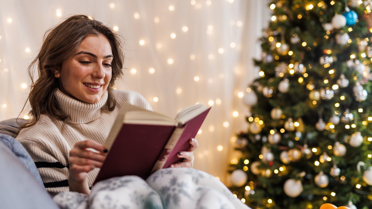Portrait of happy young woman lounging on the cozy sofa, under a warm blanket, reading an interesting book. Christmas tree and lights glistening in the background.