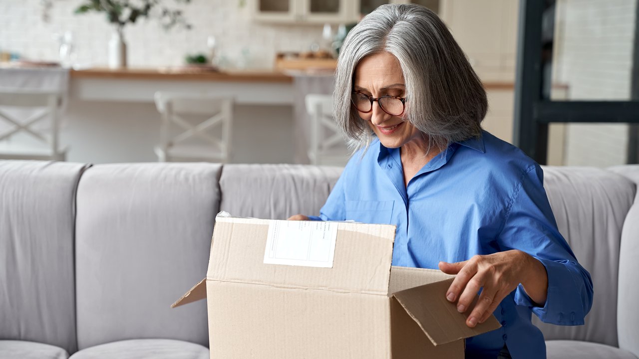 Smiling older adult mature woman customer unpacking parcel concept sitting at home on couch. Happy senior middle aged lady opening online store order receiving gift in postal delivery shipping box.