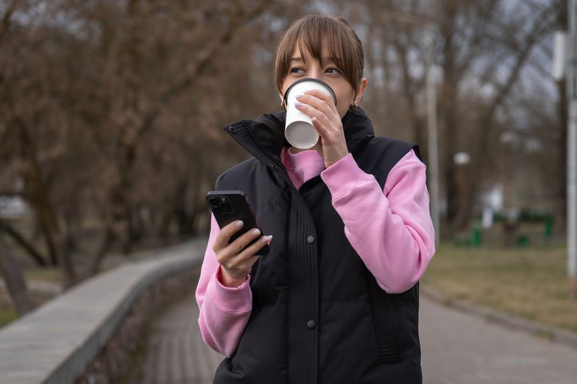 Young woman walking through a park, holding a takeaway coffee cup while browsing her smartphone, smiling at something delightful on the screen, enjoying the fresh air and nature