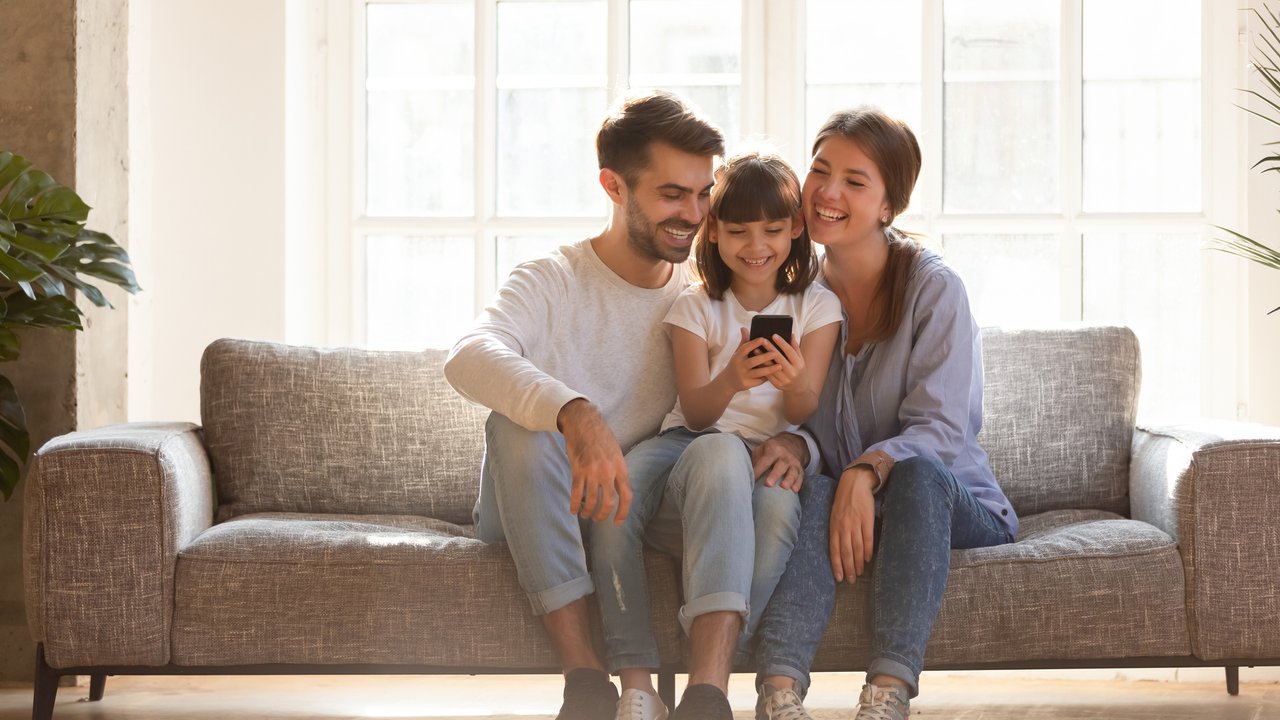 Happy family and kid daughter having fun with smartphone gadget at home, little child girl looking at phone play game using app with mom dad watching funny mobile video, making online call on couch
