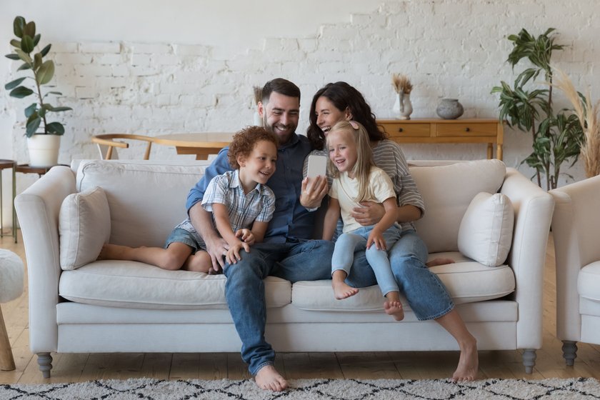 Happy excited couple of parents, little son and daughter kids talking on video call, taking selfie on cellphone, chatting, smiling, laughing, having fun, hugging on comfortable sofa at home