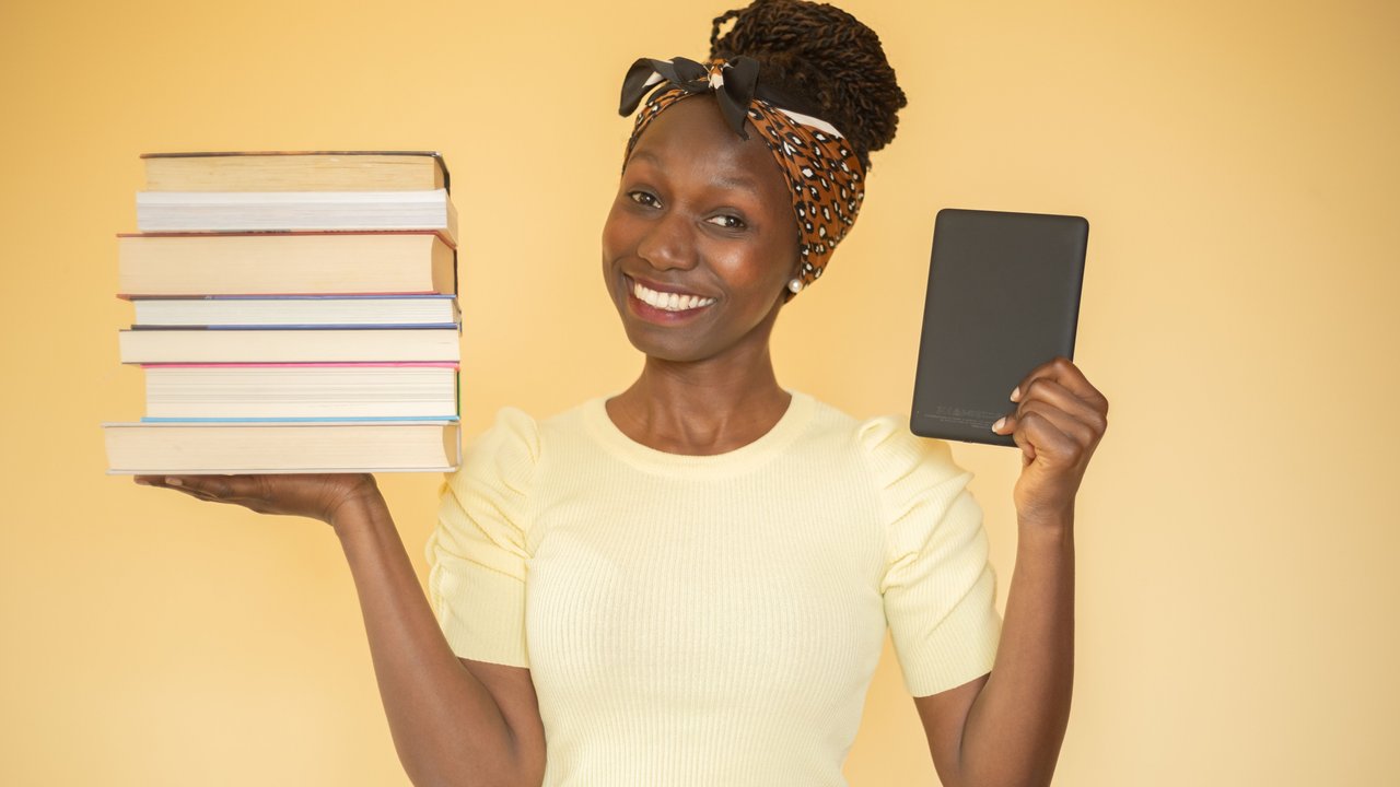 Young woman comparing printed books to e-book while carrying books on her right hand and an ebook on her left hand looking at camera