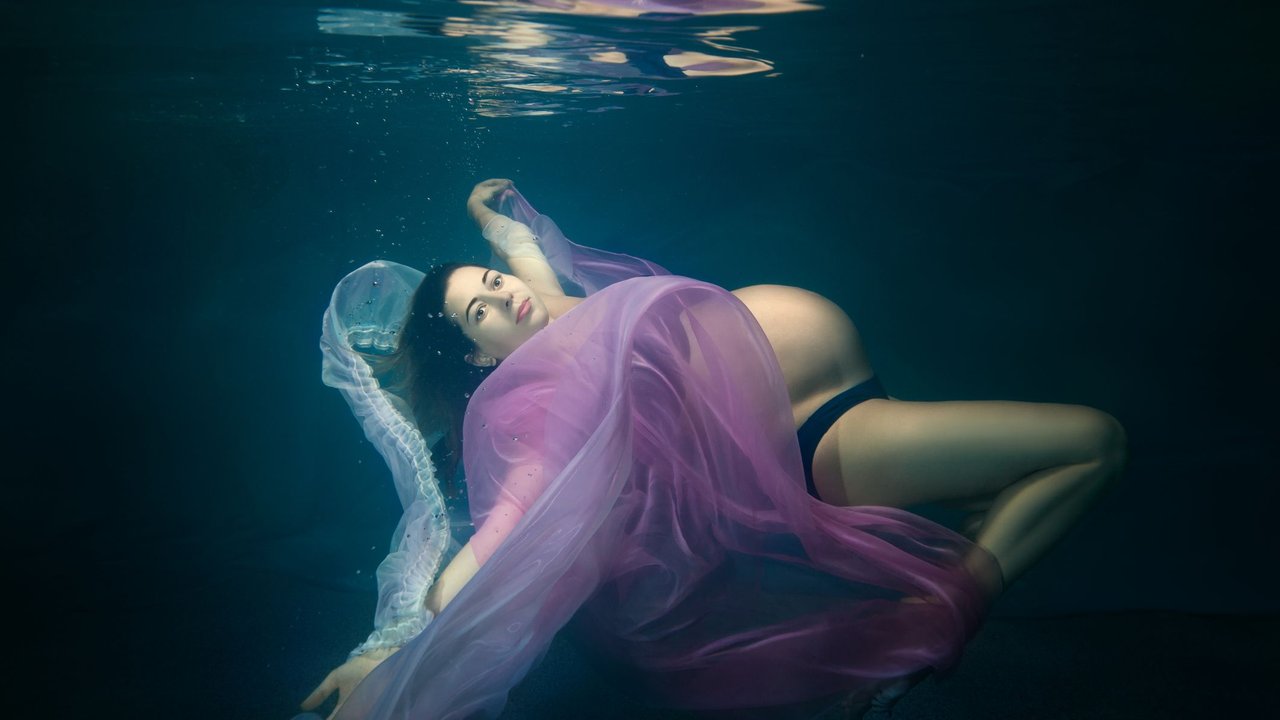 Fruchtwasser: Schwangere Frau schwimmt unter Wasser mit leichten Tüchern in blau und lila um sich herum