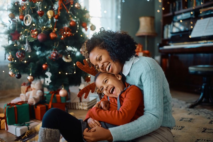 Kinder Weihnachtslieder: Mutter und Tochter sitzen lachend vor einem festlich geschmüchten Tannenbaum und lachen