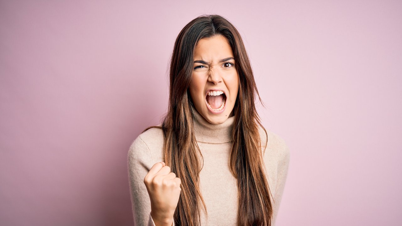 Young beautiful girl wearing casual turtleneck sweater standing over isolated pink background angry and mad raising fist frustrated and furious while shouting with anger. Rage and aggressive concept.