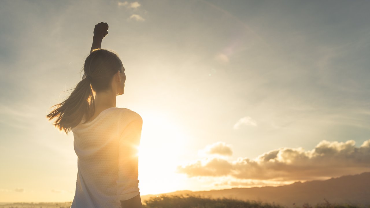 Strong, determined, confident woman with her fist up in the air facing sunset.