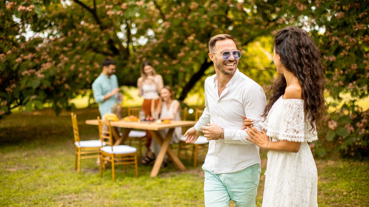 Kleidung für den Polterabend: Mann und Frau in Garten, im Hintergrund Tisch mit weiteren Personen im Sommer