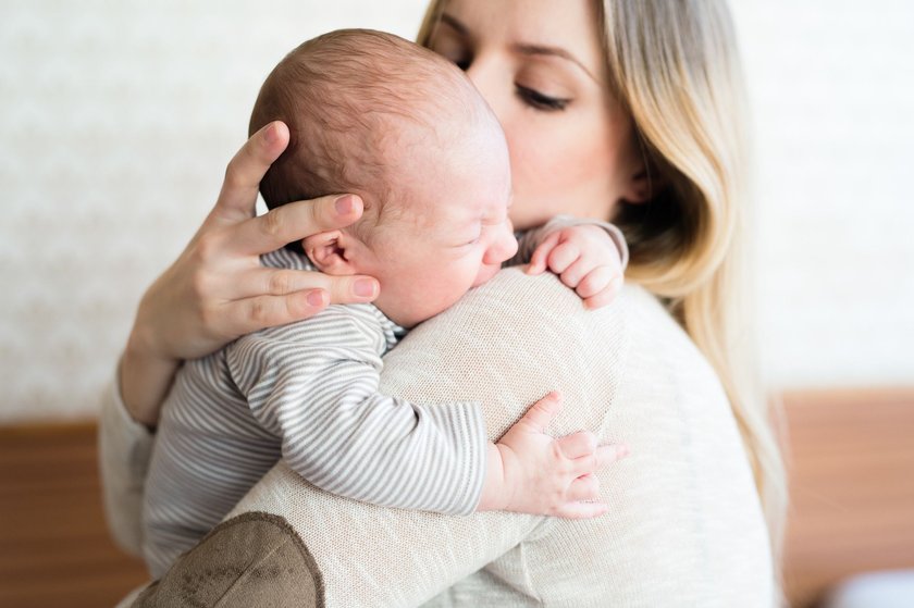 Beckenbodentraining nach der Geburt:  Blonde Mama ist zu Hause und hält weinendes Neugeborenes liebevoll tröstend an ihrer Schulter. Beide tragen helle Kleidung. 