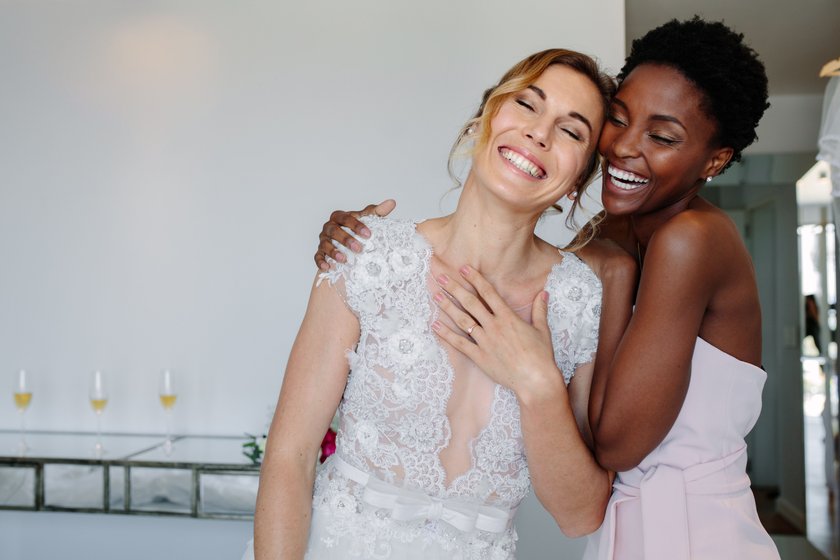 Gorgeous bride in wedding gown having fun with bridesmaid in hotel room. Cheerful bride and bridesmaid on the wedding day.