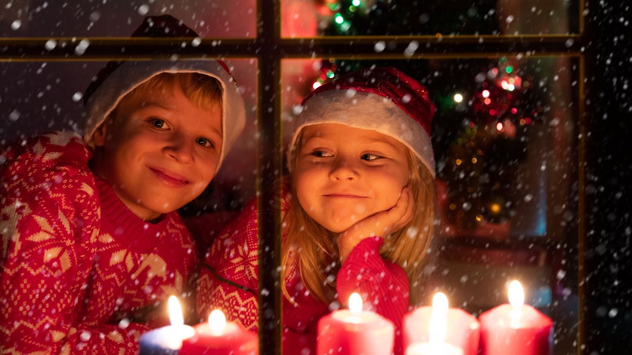 Kinder&#x20;Weihnachtslieder&#x3A;&#x20;M&#x00E4;dchen&#x20;und&#x20;Junge&#x20;in&#x20;roten&#x20;Norwegerpullis&#x20;und&#x20;mit&#x20;Nikolausm&#x00FC;tzen&#x20;schauen&#x20;durch&#x20;Fenster&#x20;mit&#x20;Kerzen&#x20;in&#x20;den&#x20;Schnee