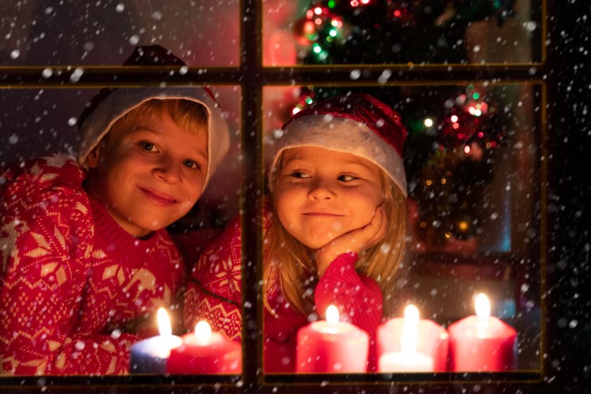 Kinder Weihnachtslieder: Mädchen und Junge in roten Norwegerpullis und mit Nikolausmützen schauen durch Fenster mit Kerzen in den Schnee