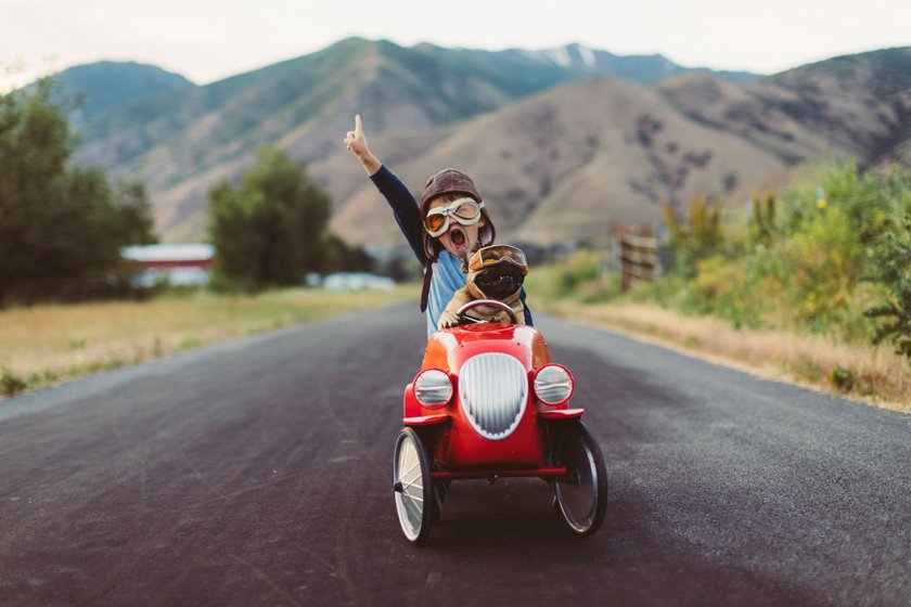 A boy wearing racing goggles races his toy race car with his support dog on a small road. This is a road trip for the soul. Image taken in Utah, USA.