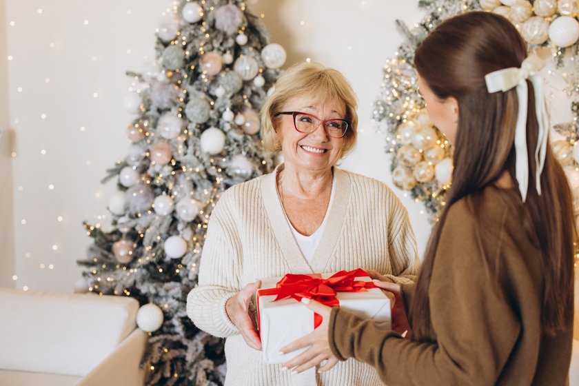A happy grandmother receives a wrapped gift from her granddaughter in beautifully decorated living room with a Christmas tree. The festive atmosphere captures the warmth and joy of the holiday season.