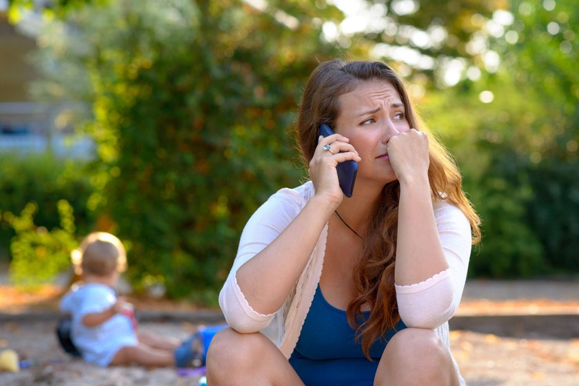 Weinende Mutter am Telefon mit spielendem Kind im Hintergrund