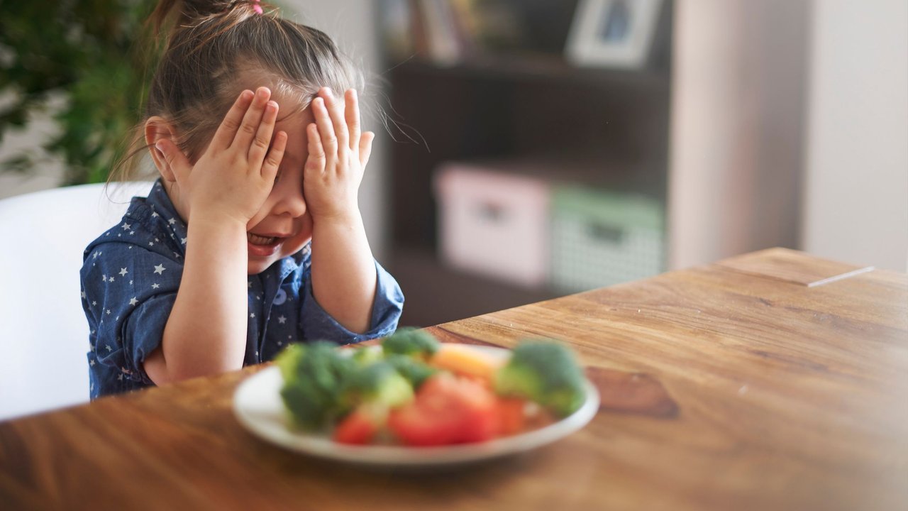 Picky Eater: Kleines Mädchen sitzt an Holzesstisch vor einem gefüllten Teller und hält sich die Augen zu