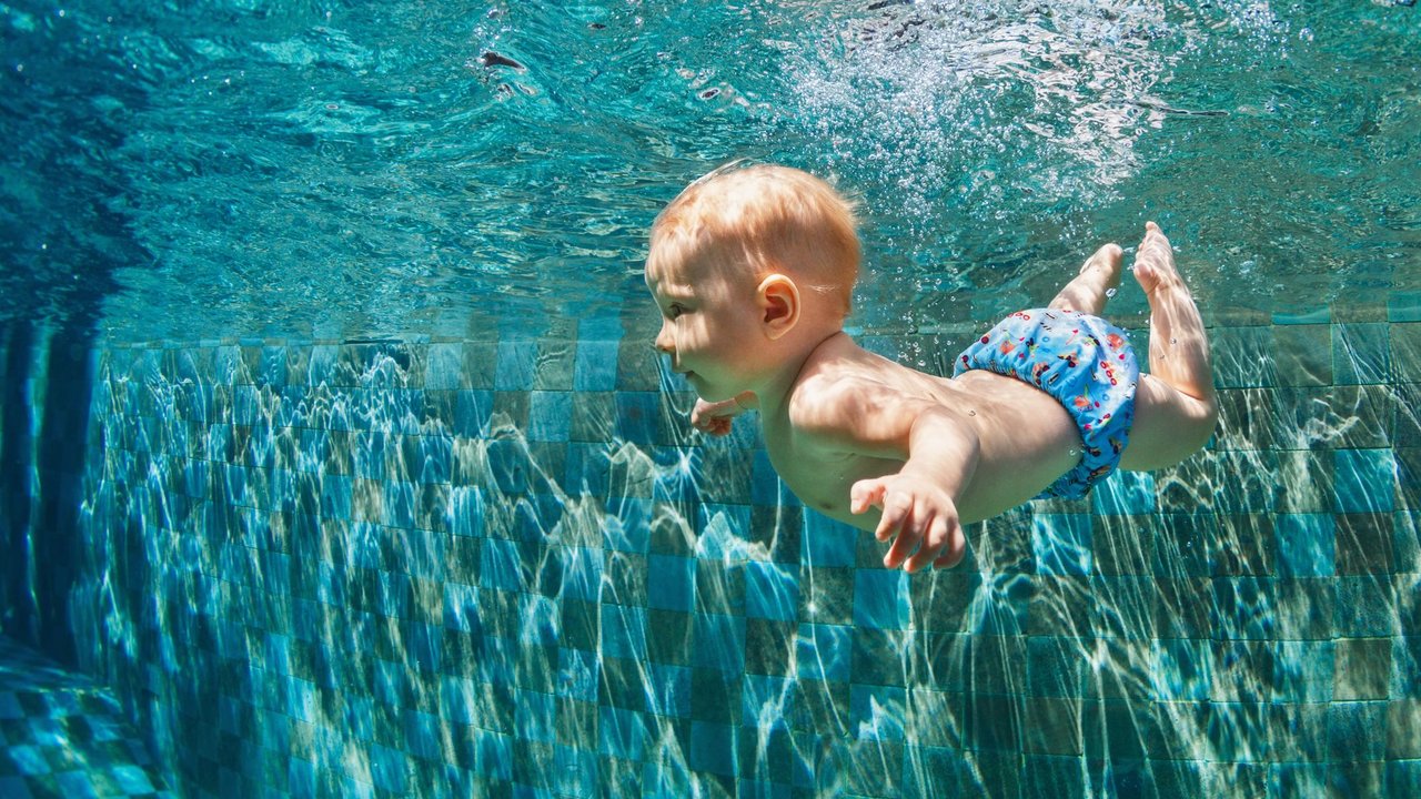 Happy child learn to swim, dive in blue pool with fun - jumping deep down underwater with splashes. Healthy family lifestyle, kids water sports activity, swimming lesson with parents in fitness club