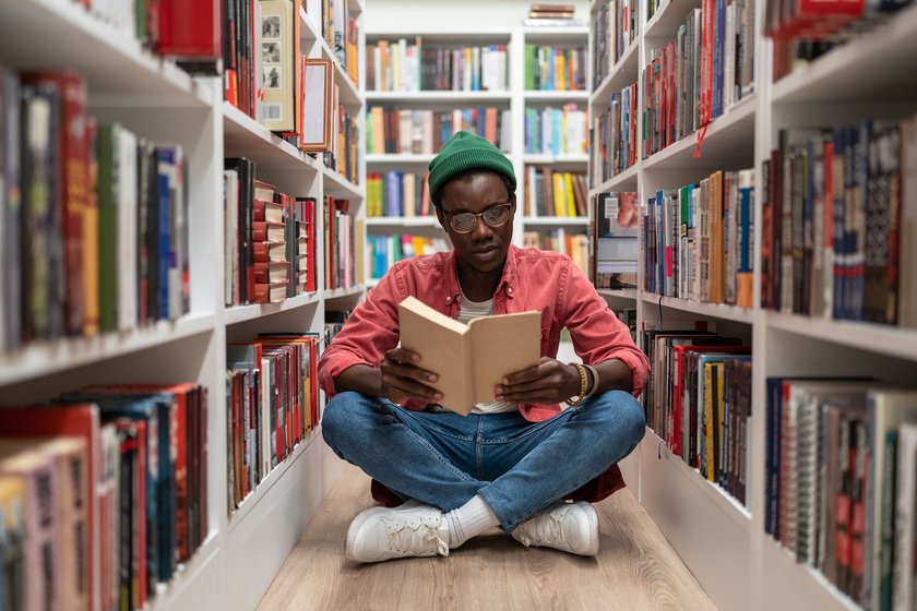 Junger Mann sitzt lesend zwischen Bücherregalen