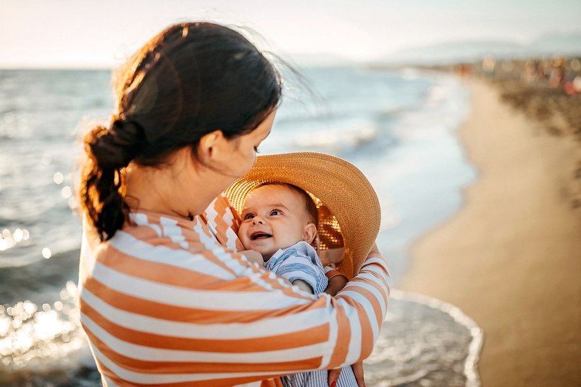 Mit dem Baby sicher durch den Sommer: Mutter schützt ihr Baby am Strand mit ihrem Strohhut vor der Sonne.
