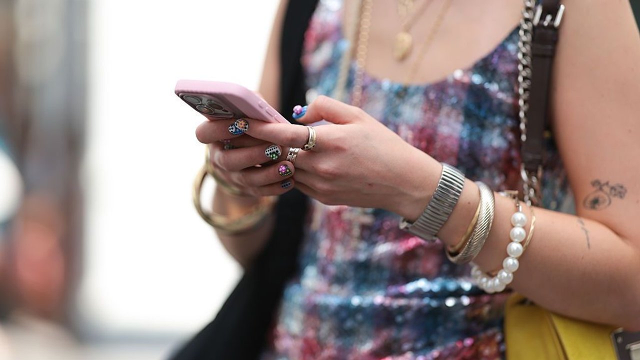 PARIS, FRANCE - JUNE 25: A Fashion Week guest is seen wearing glasses with a brown frame; several layered gold necklaces; a pink and blue striped sequin top with a round neckline; a black shoulder bag; a yellow bag with dark brown leather shoulder strap; several bangles in silver and gold and a beaded bracelet; and two rings, while holding an Apple iPhone during the Hed Mayner Menswear Spring/Summer 2026 show as part of Paris Fashion Week on June 25, 2025 in Paris, France. (Photo by Jeremy Moeller/Getty Images)