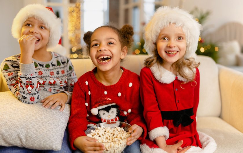 Kinder Weihnachtslieder: 3 Kinder sitzen in Weihnachtsoutfits auf Couch und essen Popkorn