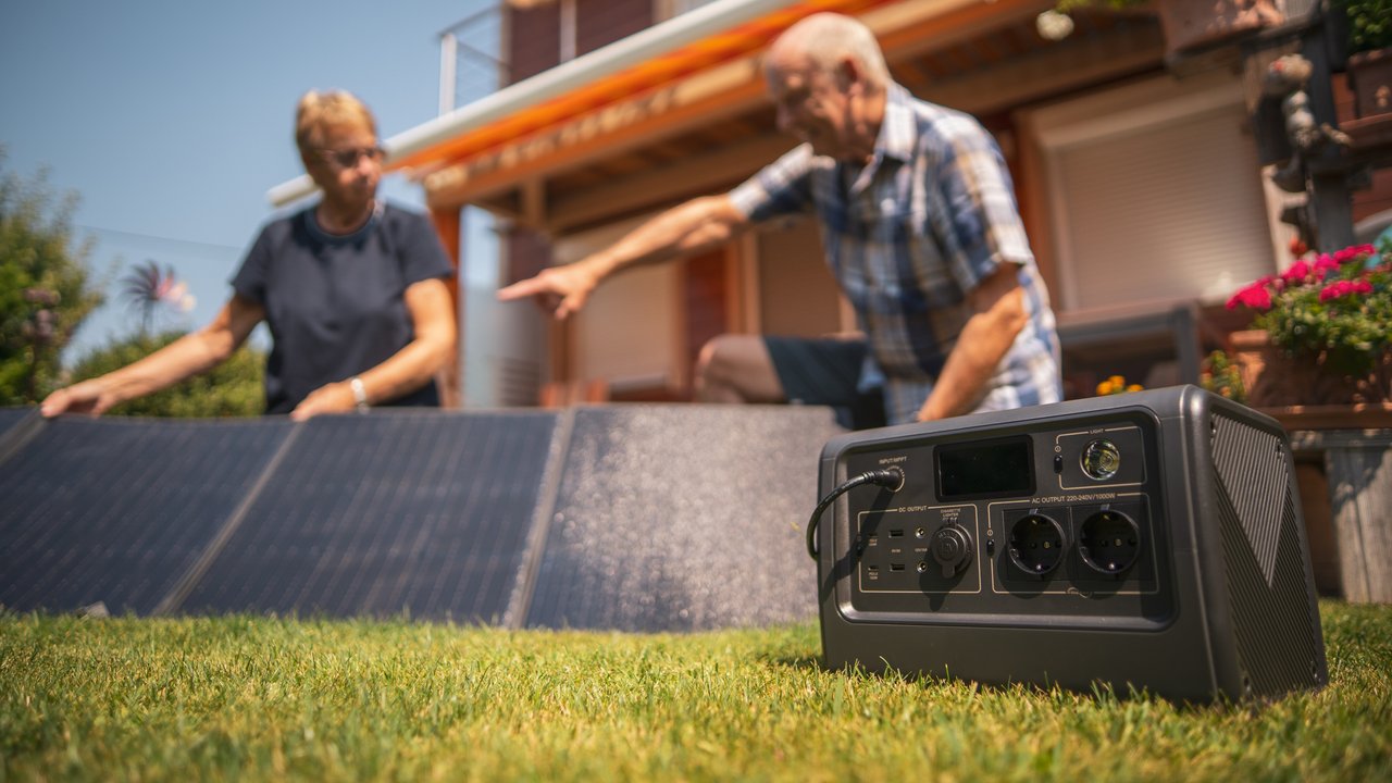 Senior couple setting up their new portable solar panels and battery on meadow in their small garden on sunny summer day, to start producing some green sun energy, focus on power back in front, background blurred