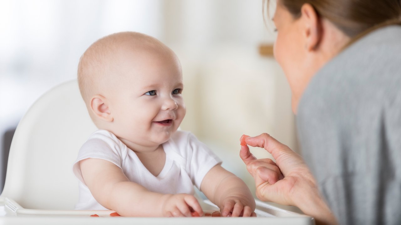 An adorable baby sits in a high chair in front of Mom and smiles at her.  She is reaching forward and offering her baby a piece of finger food.