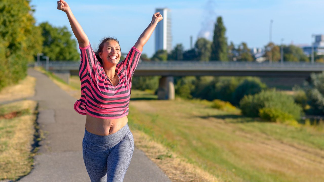 Happy fit young woman cheering and celebrating as she walks along a river after working out jogging