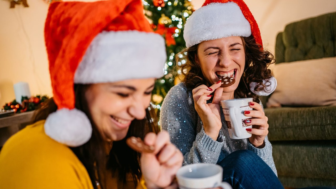 Lachende Freundinnen essen Plätzchen vor dem Weihnachtsbaum