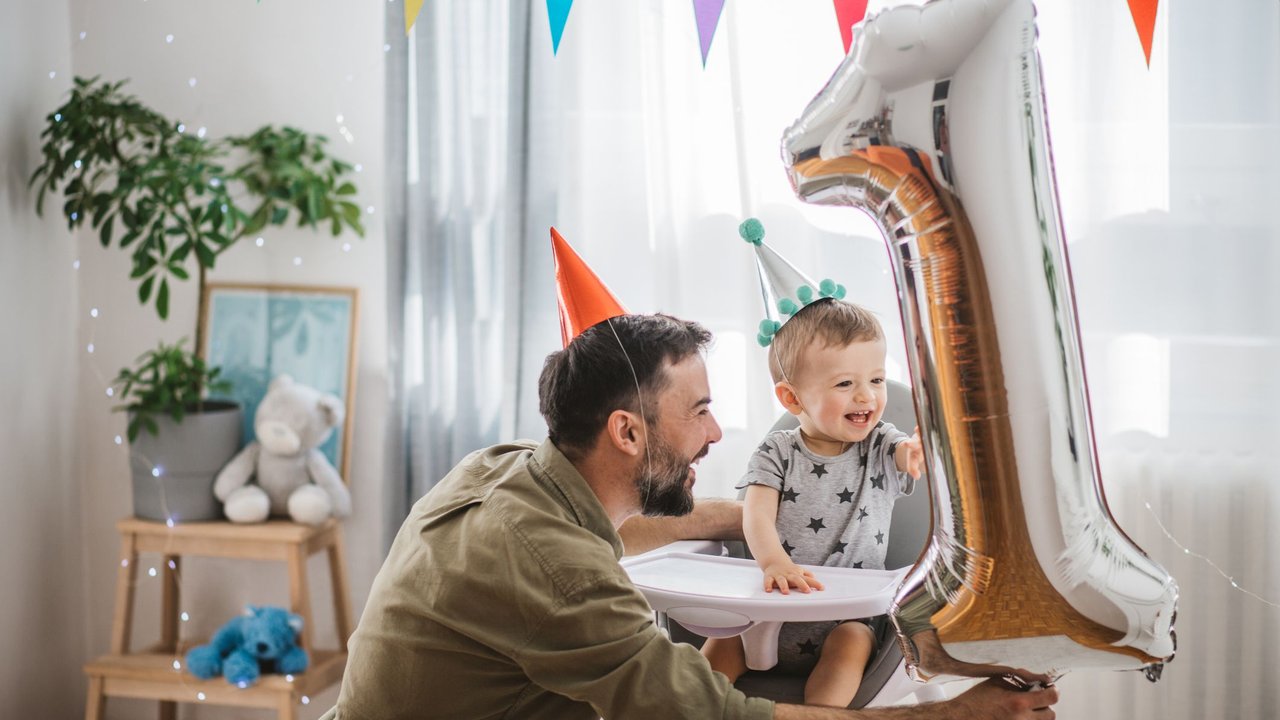 Glückwünsche zum 1. Geburtstag: Vater hält Luftballon in Form einer silbernen Eins vor sein Kind im Hochstuhl, beide tragen Partyhüte