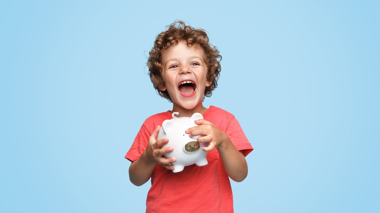Cute little boy with piggy bank laughing and looking at camera while standing on blue background
