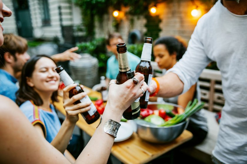 A group of friends clink beers as they make a toast at a barbecue meetup together.