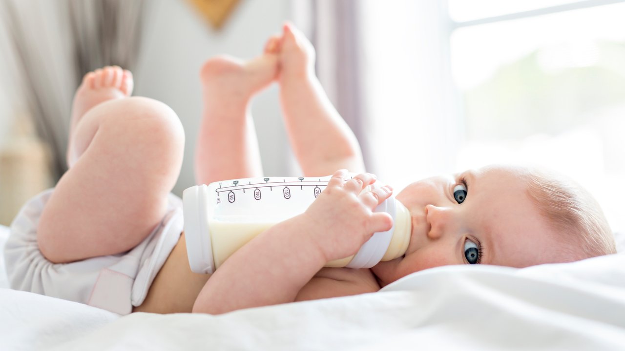 A Pretty baby girl drinks water from bottle lying on bed. Child weared diaper in nursery room.