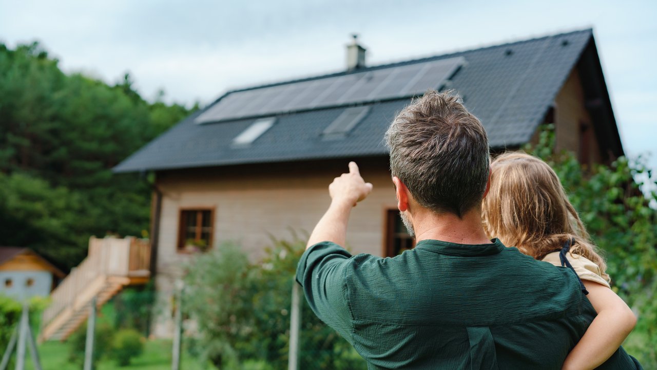Rear view of dad holding her little girl in arms and showing at their house with solar panels.Alternative energy, saving resources and sustainable lifestyle concept.