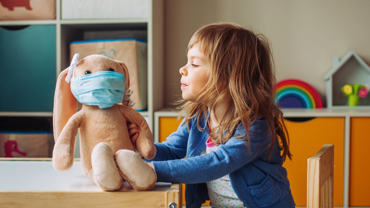 Little girl playing with rabbit soft toy in the medicine mask sitting at the table in the kids room.