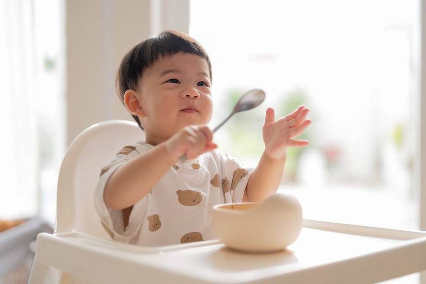 Adorable Asian baby boy eating food from the plate by himself while sitting in high chair home Baby milestone, growth and development concept