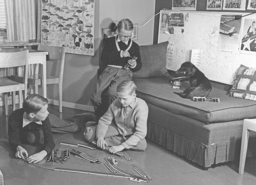 Jungen in den 1950er Jahren. Drei Jungen spielen mit einem Märklin Modelleisenbahnset. Die Teile und Gleise sind auf dem Boden ausgelegt und werden mit Strom betrieben.