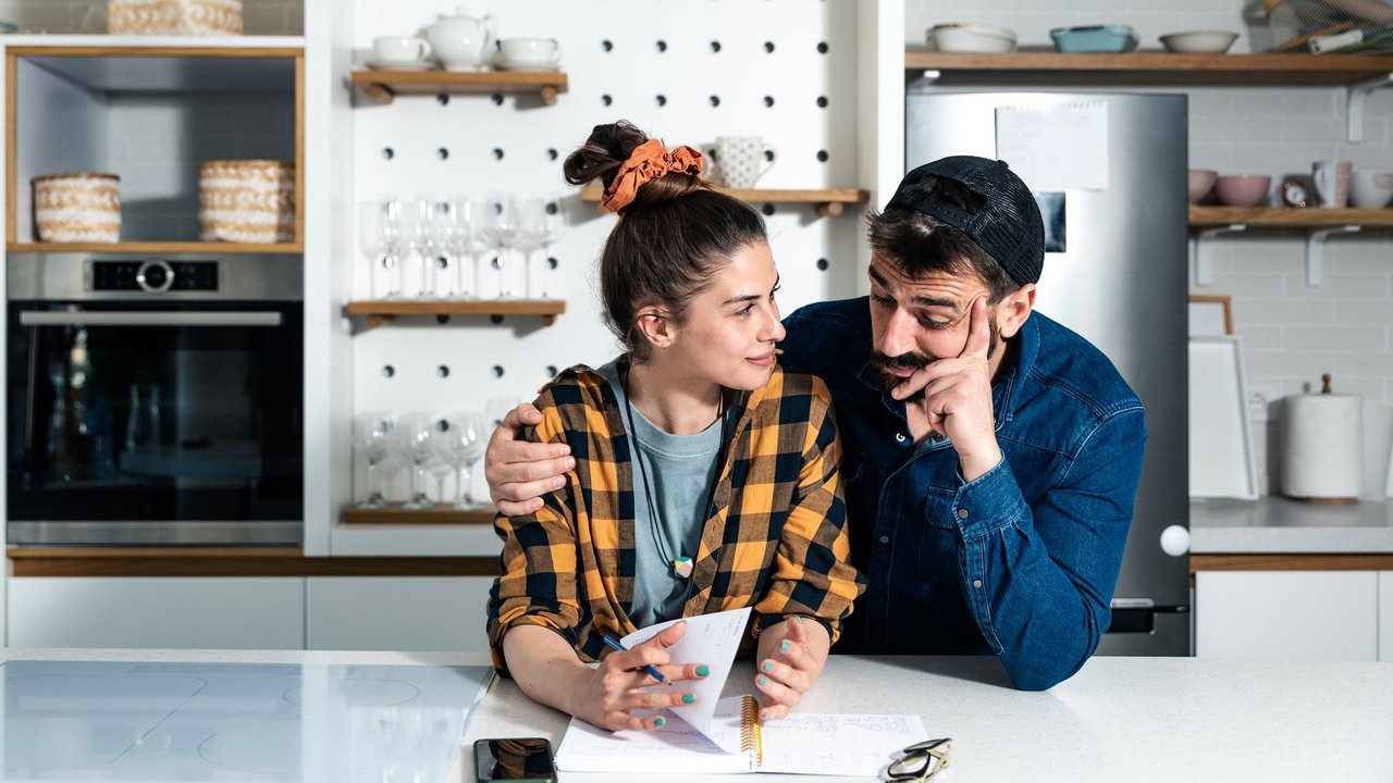Einladung für Polterabend: Mann und Frau sitzen in Küche vor einem Block mit Stift und Handy und schauen nachdenklich