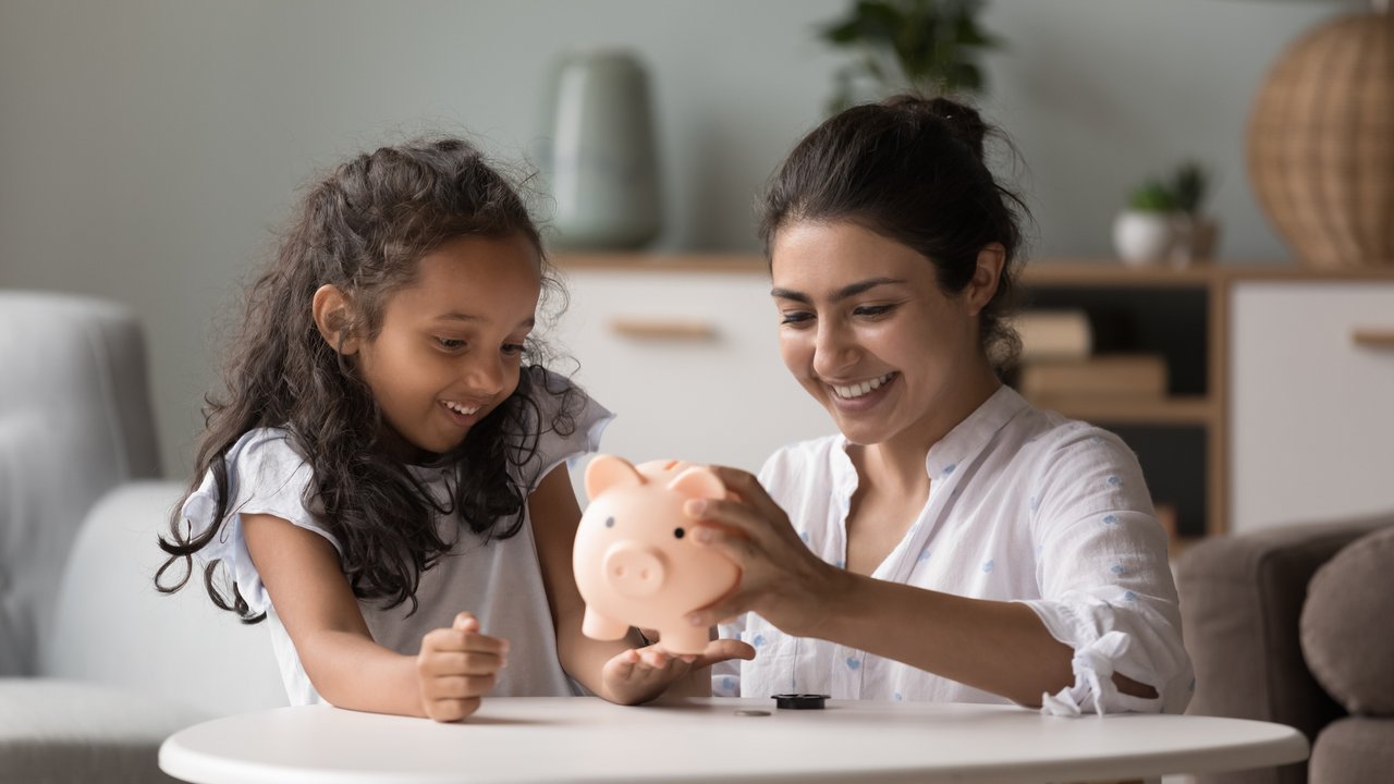 Young happy Indian woman and preschooler daughter sitting at table in living room take out money stack from piggy bank for spending money for future purchases, smile enjoy moment. Savings, finances