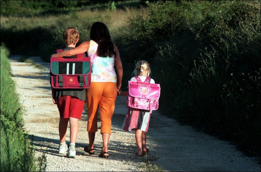 Eine Mutter geht mit zwei Kindern, die einen Schulranzen auf dem Rücken tragen, einen Weg entlang